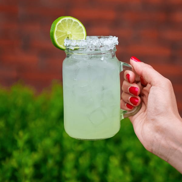 A hand with red nails holds a Tacos & Bla Bla Bla margarita in a mason jar with a lime wedge.