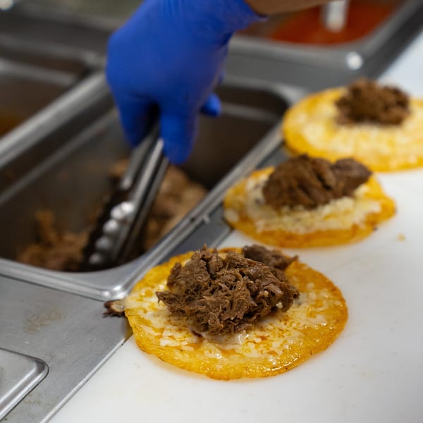 A gloved hand uses tongs to add shredded meat to a cheesy tortilla, preparing Tacos & Bla Bla Bla tacos.