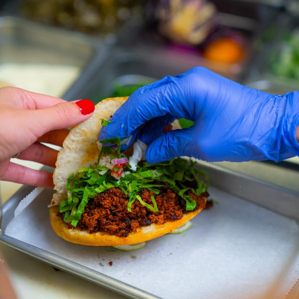 Hands assembling a Mexican torta with seasoned meat, lettuce, and pico de gallo.