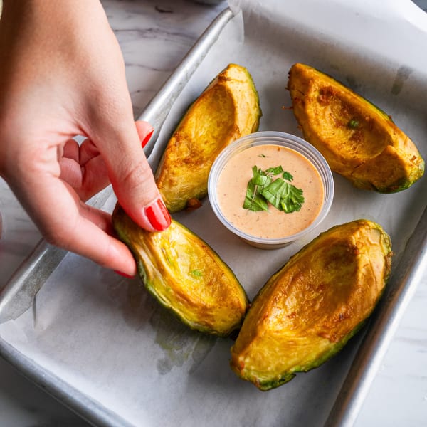 A hand placing a golden-brown avocado half next to a dipping sauce on a baking sheet.