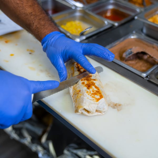 Gloved hands slicing a grilled burrito on a white cutting board, with food pans in the background.