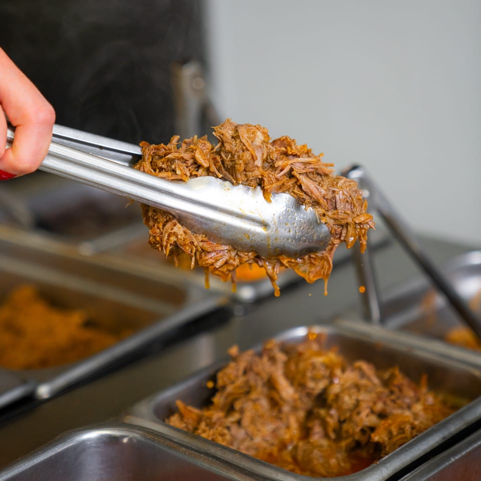 Tongs lifting juicy, shredded Mexican barbacoa from a steaming buffet tray at Tacos & Bla Bla Bla.