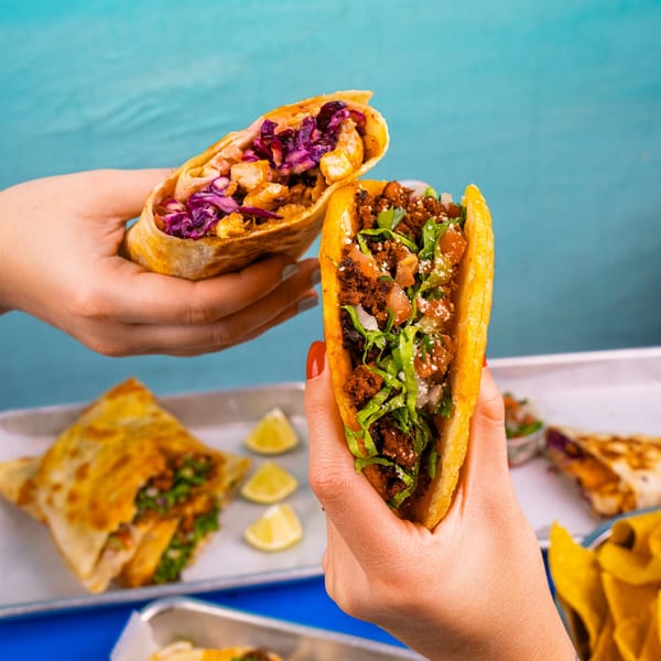 Hands holding a vibrant taco and a burrito, with more Mexican food on trays below.