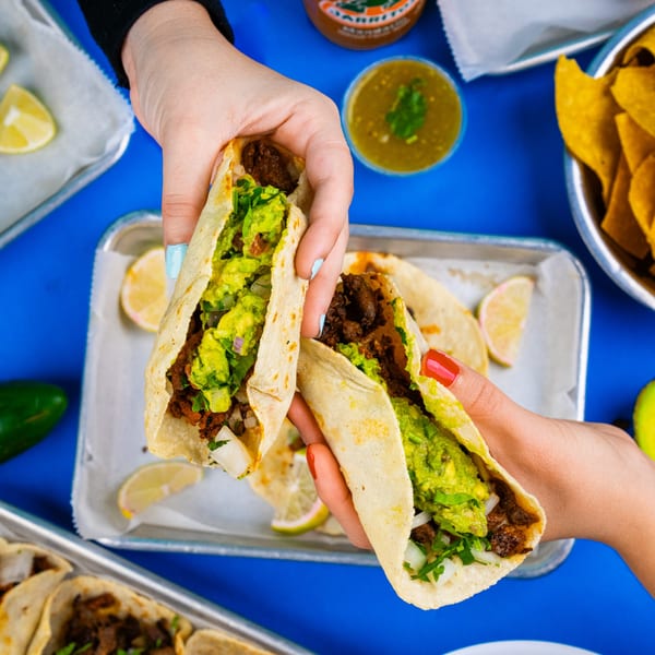 Two hands holding up Tacos & Bla Bla Bla tacos filled with meat, guacamole, and onions.
