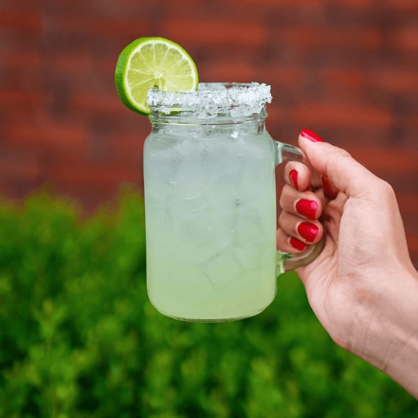 A hand with red nails holds a Tacos & Bla Bla Bla margarita with a salted rim and lime.