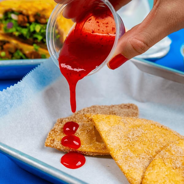 Hand pouring red strawberry sauce from a plastic cup onto two sugar-dusted Mexican dessert empanadas.