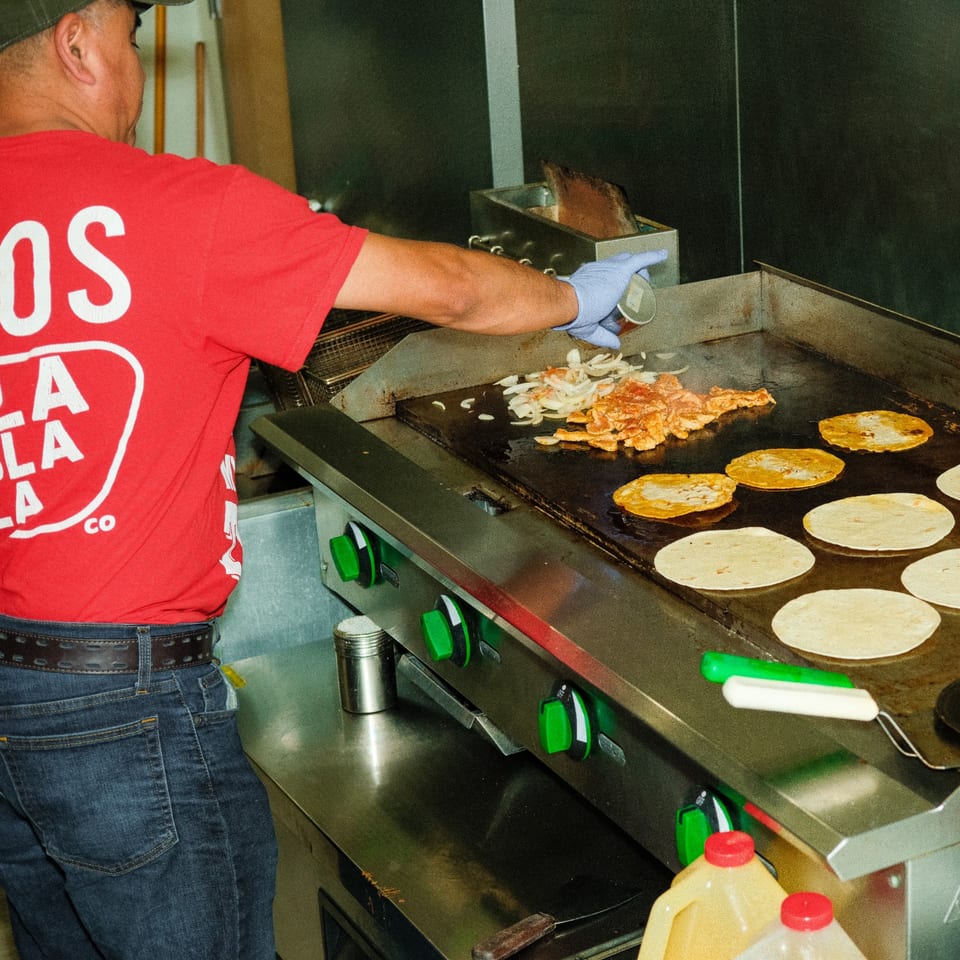 A chef in a red Tacos & Bla Bla Bla shirt cooks tortillas, meat, and onions on a griddle.