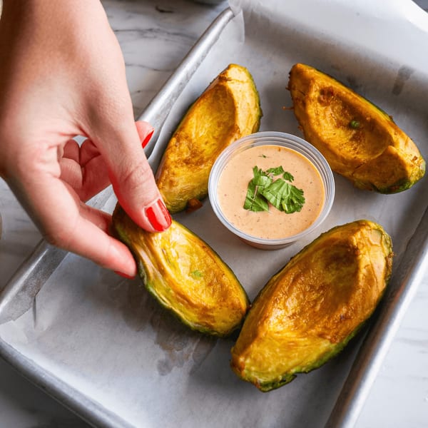 A hand with red nail polish reaching for a baked avocado half with a side of sauce.