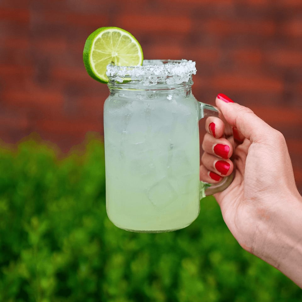 A hand with red nails holds a Tacos & Bla Bla Bla margarita in a mason jar with a lime slice.