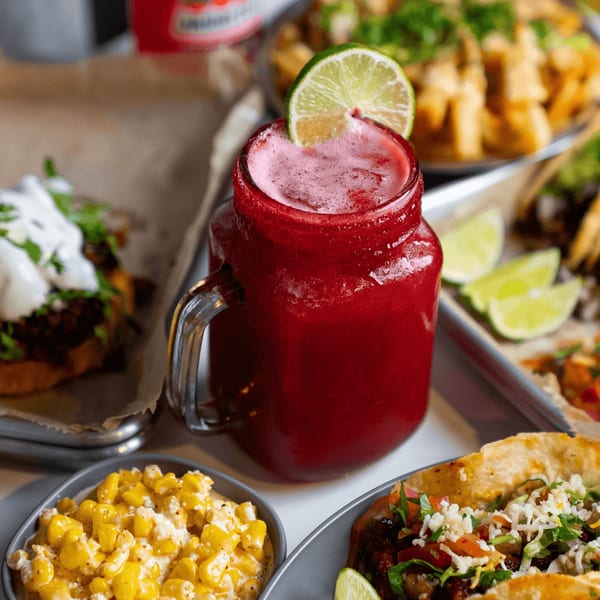 A vibrant red drink with a lime wedge, surrounded by Mexican dishes like tacos and elote.