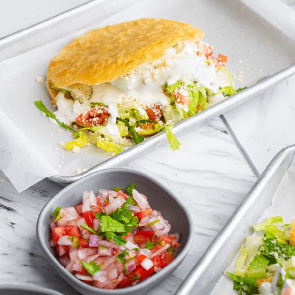 A golden-fried gordita filled with lettuce, tomatoes, and white sauce, next to a bowl of pico de gallo.
