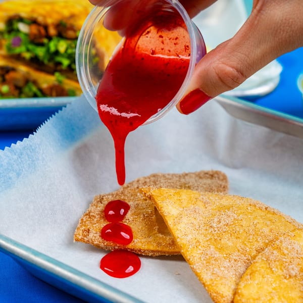 Hand pouring red strawberry sauce from a plastic cup onto two sugar-dusted Mexican dessert pastries.