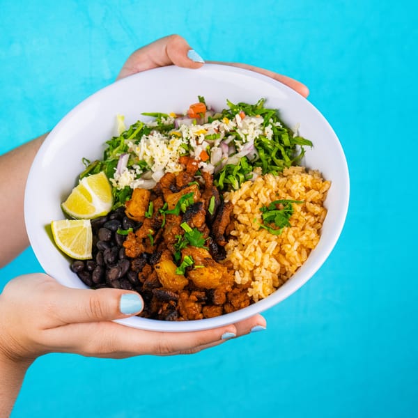 Hands holding a Mexican bowl with rice, beans, meat, salad, cheese, and lime wedges.