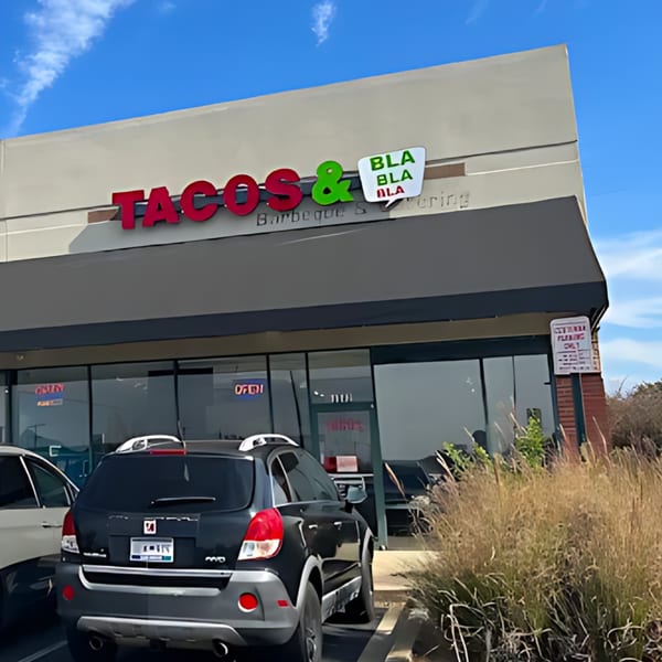 Exterior of Tacos & Bla Bla Bla restaurant with its sign, parked cars, and dry grass in the foreground.
